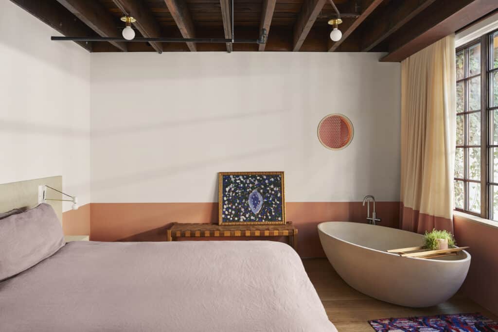 A modern bedroom with exposed wooden ceiling beams, a bed with lavender bedding, a freestanding oval bathtub, a wooden bench with a framed floral picture, and large windows with beige curtains.