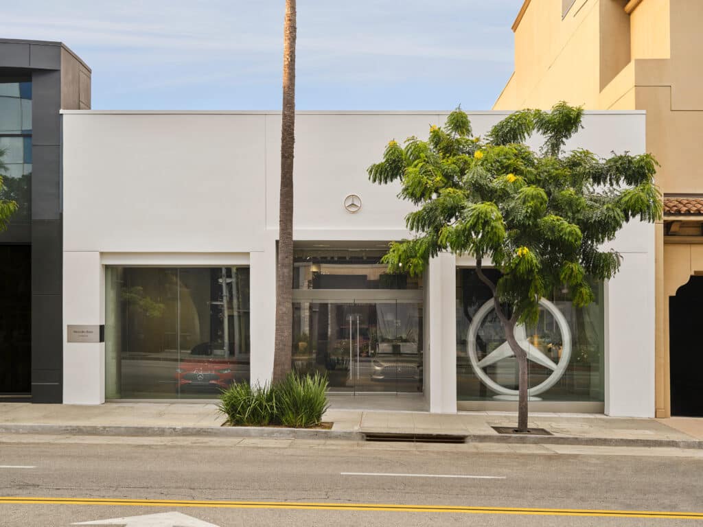 A modern white building with large glass windows, a Mercedes-Benz logo above the entrance, and a tree on the sidewalk in front. A palm tree is also visible behind the building.