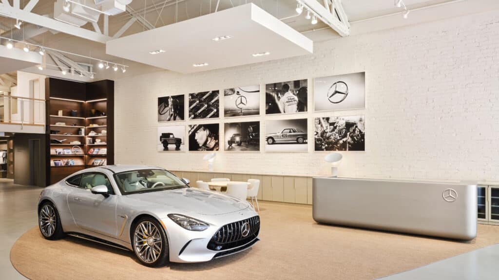 A silver Mercedes-Benz sports car is displayed in a modern showroom with white walls, a reception desk, and black-and-white photos of Mercedes vehicles and logos on the wall. Bright lighting and shelves with books are visible in the background.