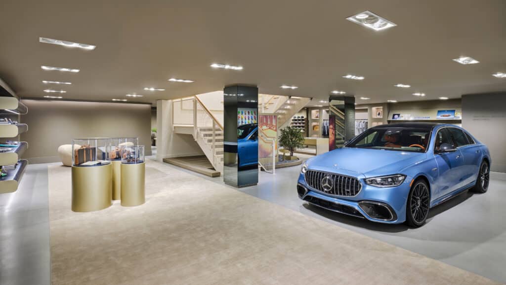 A modern showroom with a blue luxury car, designer handbags in a glass display, shelves with accessories, and a staircase in the background. The space is brightly lit and elegant.