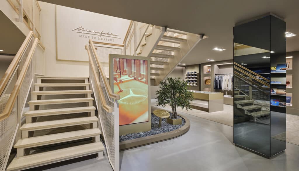 Modern clothing store interior with a staircase, digital display, potted plants, shelves with folded clothes, and a large mirror. A sign reading “Made to Measure” hangs on the wall above the stairs.