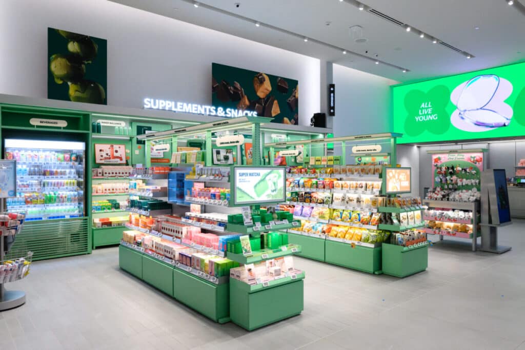 A brightly lit, modern store interior features green shelves stocked with supplements, snacks, and wellness products. Refrigerated drinks and snacks are displayed on the left. Digital signs and product displays fill the space.