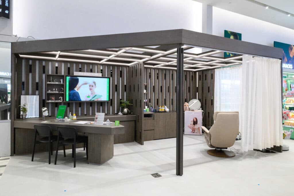Modern consultation booth with dark wood decor, chairs, a desk, TV screen, and beauty products. A privacy curtain separates a reclining chair. The setting appears to be in a shopping mall or retail space.
