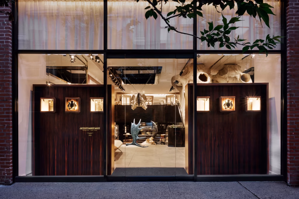 Street view of a modern store with large glass windows, featuring dark wooden doors, illuminated display boxes, and a sculptural object visible inside the well-lit interior.