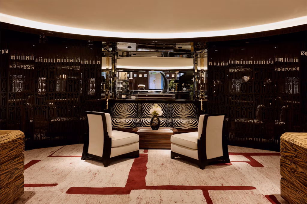 Modern hotel lounge with two white armchairs facing a small table with a flower vase. Geometric wall panels, patterned carpet, and a sleek bar area are visible in the background. Warm lighting creates an inviting atmosphere.
