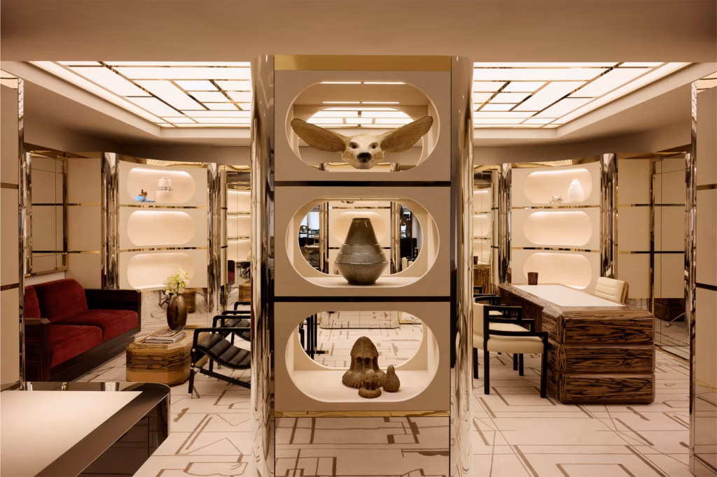 Modern, luxurious boutique interior with gold accents, open shelves displaying art pieces, a wood desk with a white chair, a red sofa, black chairs, and geometric lighting on the ceiling and patterned floor.